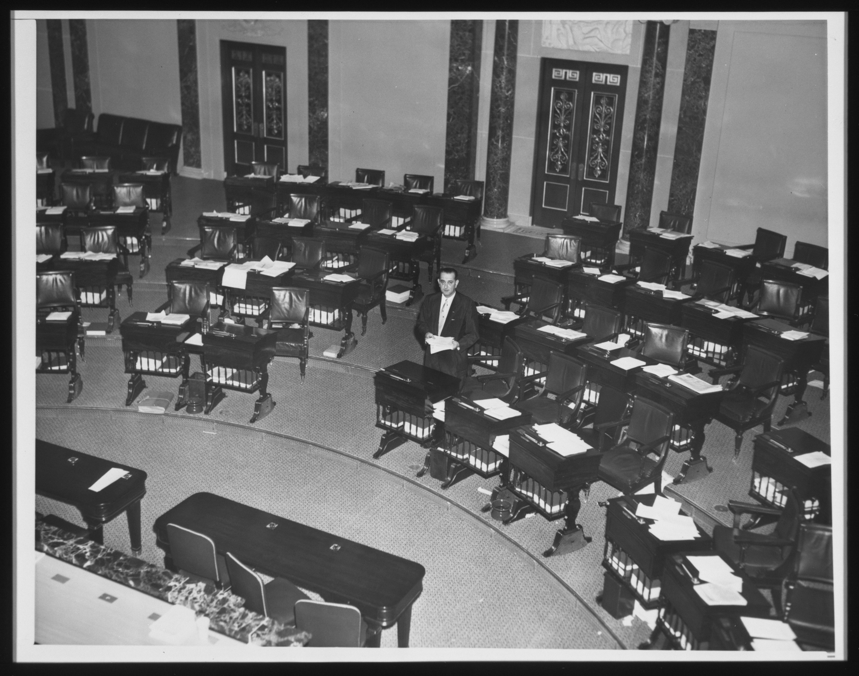A wide shot from above. Senator Lyndon B. Johnson, alone in the senate chamber, cleans out his desk. 